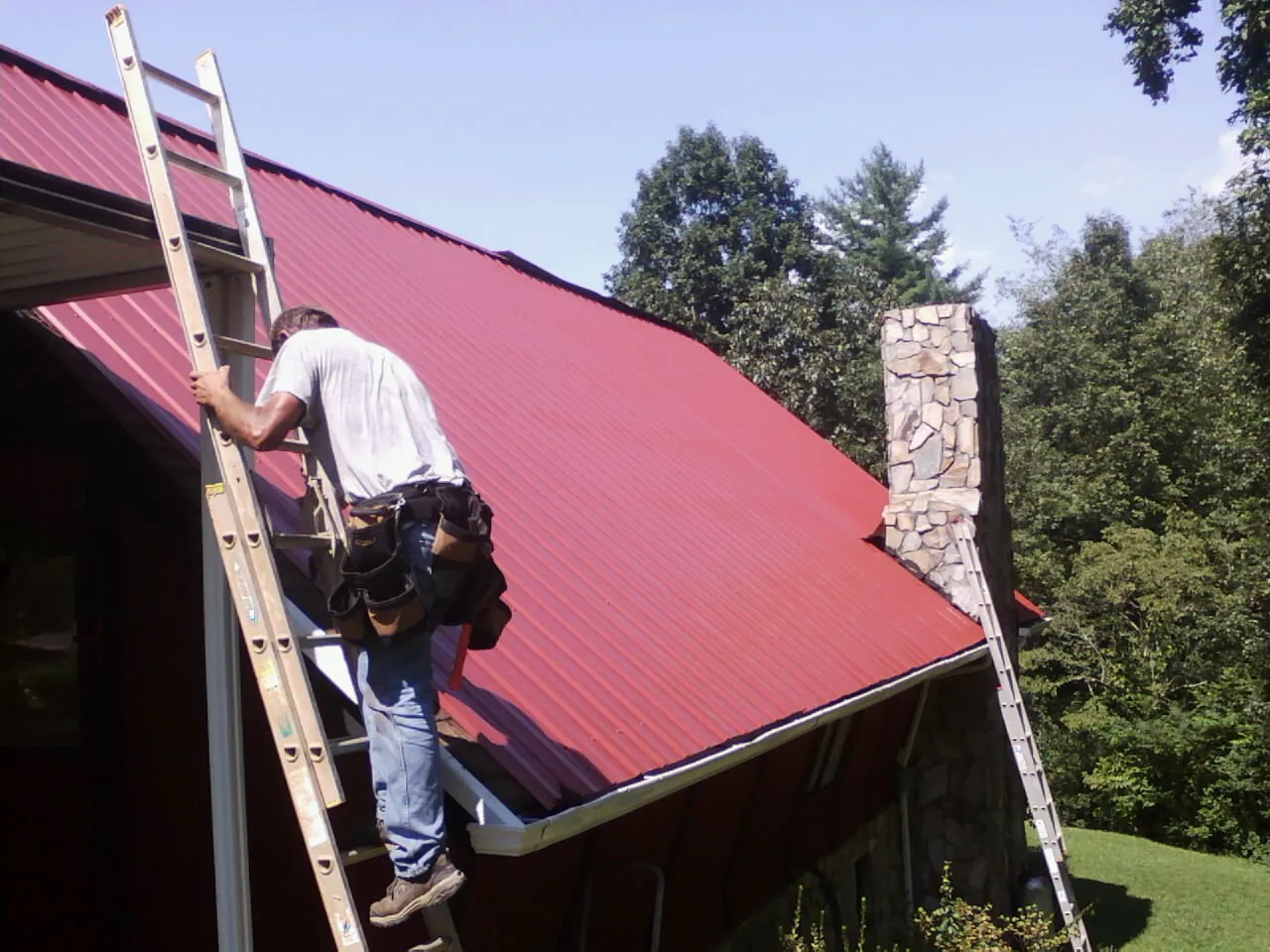 Roofer on a ladder beside a red metal roof and stone chimney — Soffit & Fascia Repair by Roofing Brothers in Warsaw